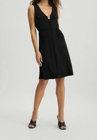 Musta hihaton mekko V-pääntiellä, sidottu vyötäröllä ja kullanvärisellä yksityiskohdalla rinnassa. Pehmeä kangas, lörppösilhouette ja polvipituinen leikkaus.