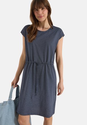 Młoda kobieta ubrana w niebieską sukienkę w paski z krótkim rękawem i wiązaniem w talii, trzyma dużą jasnoniebieską torbę na ramię, uśmiecha się do aparatu.