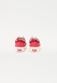 Red suede shoes with a low-top design, white rubber soles, and a red logo on the heel. Features contrasting stitching and a smooth texture.