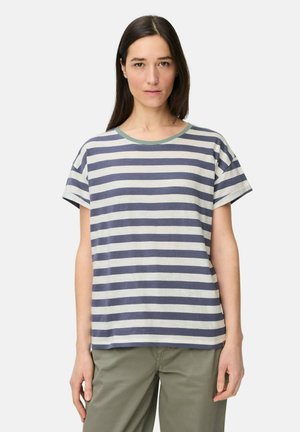 Kvinde med langt mørkt hår iført en kortærmet, blå og hvid stribet T-shirt og olivengrønne bukser, stående mod en ensfarvet baggrund.