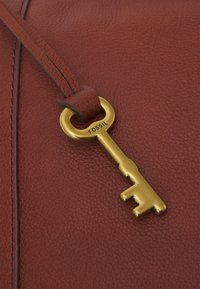 Brun lædertaske med en tekstureret overflade, der har en fremtrædende messingnøglecharme mærket "FOSSIL" fastgjort til en matchende rem.