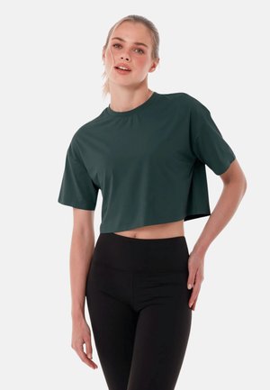 Młoda kobieta o jasnej skórze i blond włosach nosi ciemnozielony, krótki t-shirt oraz czarne legginsy z wysokim stanem, stoi na jasnym tle.