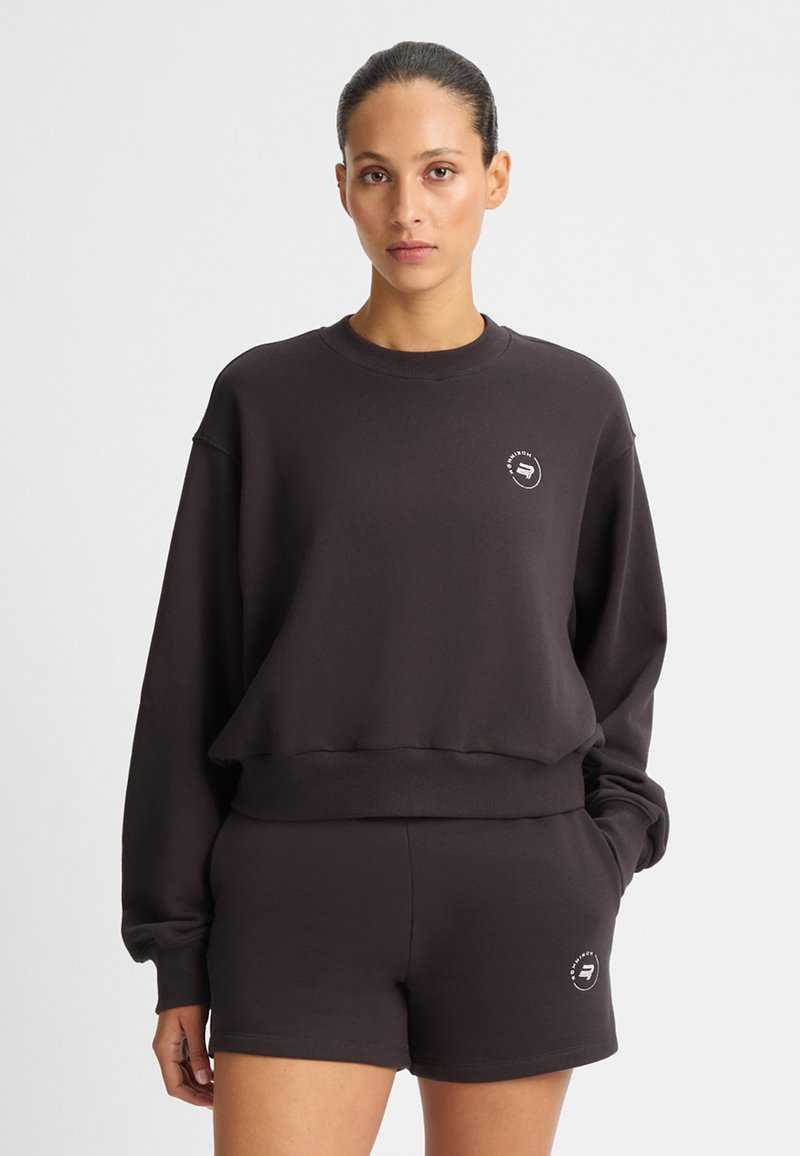 Completo con felpa nera e pantaloni corti, realizzato in tessuto morbido. La felpa ha un colletto tondo e un dettaglio del logo sul petto. Vestibilità casual e rilassata.