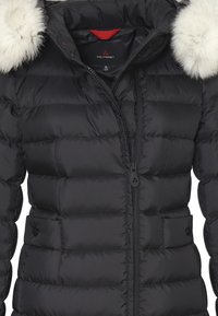 Giacca a vento nera con cappuccio foderato in pelliccia. Presenta una texture trapuntata, chiusura a doppia zip e un accento rosso dietro il colletto.