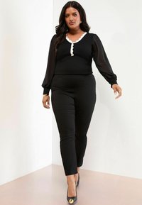 Sort tætsiddende jumpsuit med ribbet tekstur og gennemsigtige puffærmer. Har en v-hals med bølget hvid kant og knapdetaljer.