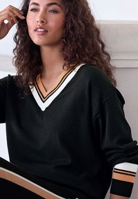 Schwarzer Pullover mit V-Ausschnitt, der weiße, orange und schwarze Streifen am Halsbündchen und an den Bündchen aufweist. Weicher Textur und lässiger Schnitt.