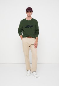 Młody mężczyzna ubrany w zielony sweter Hilfiger, beżowe spodnie, brązowy pasek, białe trampki i ciemnoczerwoną czapkę baseballową, stojący z rękami w kieszeniach.