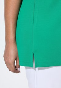 Top in cotone verde con fessure laterali, caratterizzato da una trama liscia e dettagli di cucitura sull'orlo, abbinato a pantaloni bianchi.