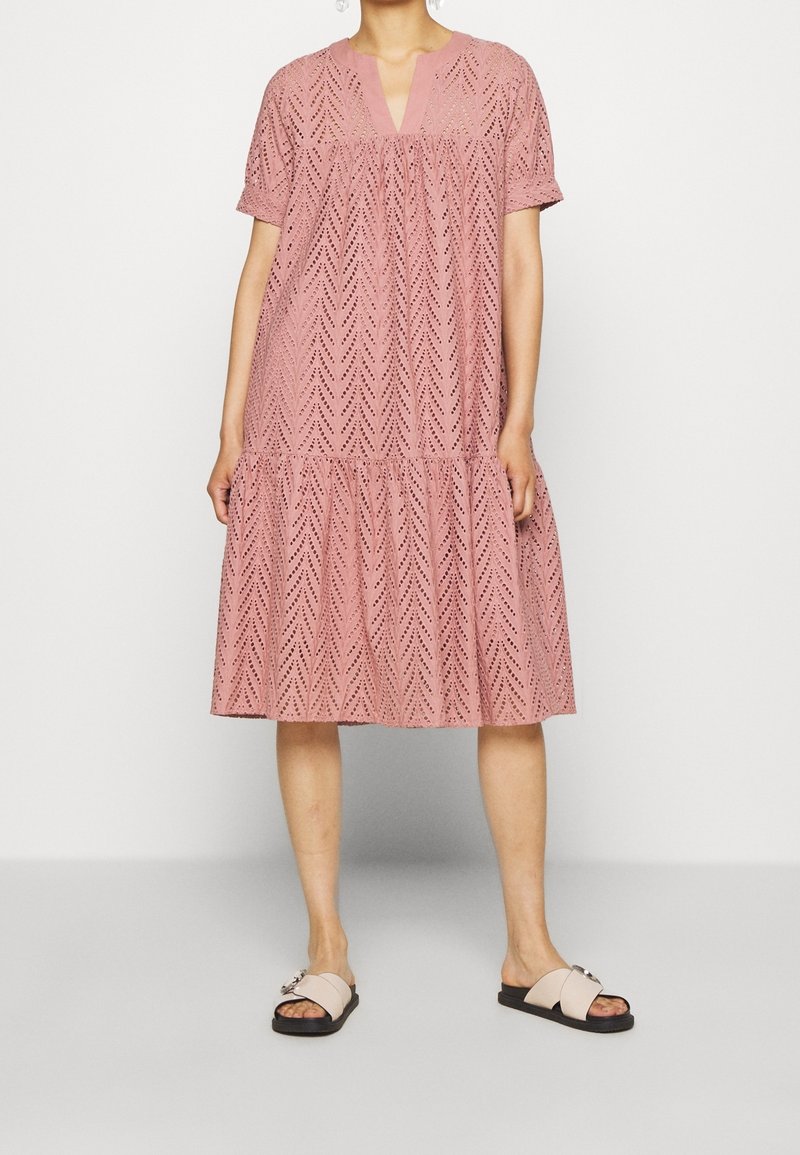 Robe rose pâle à manches courtes, col en V, motif ajouré et design à étages, associée à des sandales légères avec un détail boucle.