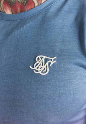 Nærbillede af en blå skjorte, der viser hvide broderede overlappende initialer og en del af et rødt og sort brysttatovering ved halsudskæringen.