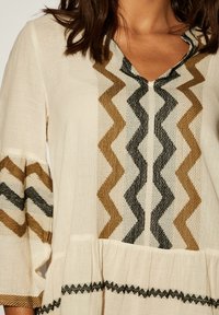 Tunique crème avec des motifs en zigzag en noir, marron foncé et or. Tissu doux avec un décolleté en V et des manches évasées, détaillées au niveau de l'ourlet.