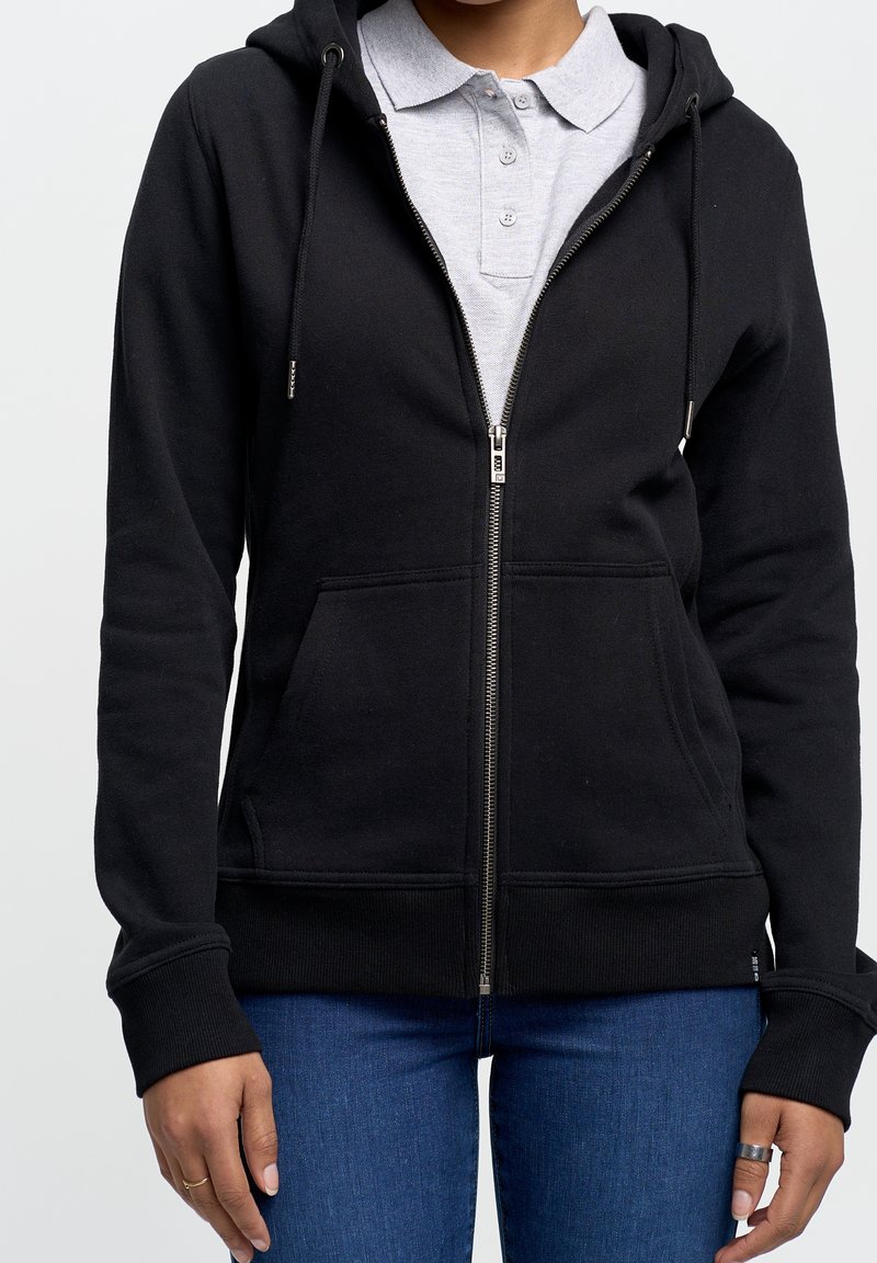 Felpa nera con zip realizzata in tessuto morbido, dotata di tasca anteriore, cappuccio con coulisse e polsini a costine, indossata sopra una camicia grigia con colletto.
