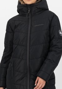 Giubbotto di piumino nero con cappuccio aderente, chiusura frontale con zip e tasche laterali. Caratterizzato da una texture trapuntata e un logo sulla manica.