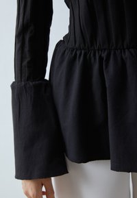 Svart langermet topp med en tettsittende overdel, samlet midje og utsvungne ermer. Stoffet virker mykt med en jevn tekstur.