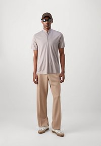 Muž v béžové košili s krátkým rukávem, volných béžových kalhotách, bílých a hnědých teniskách, tmavých slunečních brýlích a tmavé čepici, stojí proti jednobarevnému pozadí.