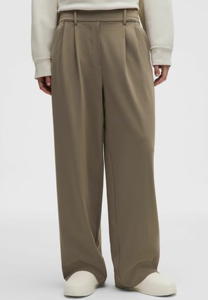 Person iført beige bukser med vide ben og hvide slip-on sko, hænder afslappet langs siden, stående på ensartet lys baggrund.