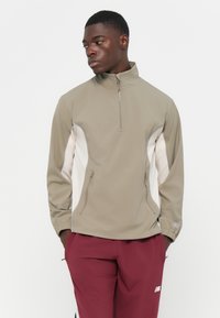 Letvægts halv-zip jakke i lysebrun med hvide sidelapper. Har to lynlåslommer foran og elastiske manchetter. Glat, mat tekstur.
