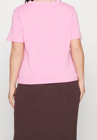 Ljusrosa kortärmad t-shirt med vågig halsringning, tillverkad av mjukt bomullsmaterial, i kombination med en brun ribbad kjol.