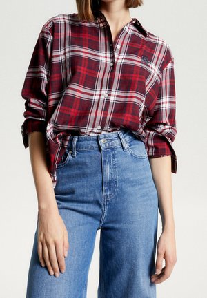 Femme portant une chemise à carreaux rouge à boutons, manches retroussées, rentrée dans un jean taille haute bleu, debout devant un fond uni.