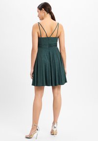 Abito verde brillante con corpino aderente e gonna svasata. Presenta spalline sottili, vita arricciata e schiena semplice. Indossato con tacchi argento.