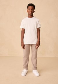 T-shirt en coton blanc, pantalon de survêtement beige et baskets blanches. Le mannequin se tient devant un fond beige clair, les bras détendus le long du corps.