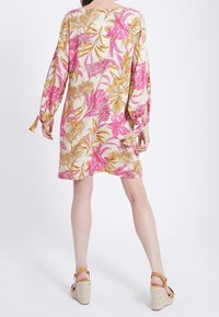 Blommig tunikaklänning med långa ärmar, med en beige bakgrund, rosa och gula botaniska mönster, och knutna manschetter. Bärs med kilklacksandaler.