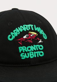 Gorra de béisbol negra con gráficos bordados coloridos: personaje de dibujos animados rojo con grandes ojos, texto "CARHARTT WIP" y "PRONTO SUBITO" en verde.