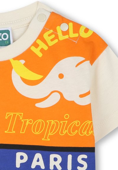 La t-shirt in cotone a maniche corte presenta colori arancione e crema, una grafica di un elefante, testo giallo, chiusure a pressione sulla spalla e motivi audaci.