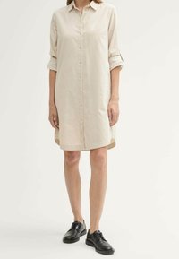 Abito camicia beige con bottoni e maniche lunghe arrotolate ai polsi. Presenta una linea dritta e un orlo posteriore leggermente più lungo. Indossato con scarpe nere.