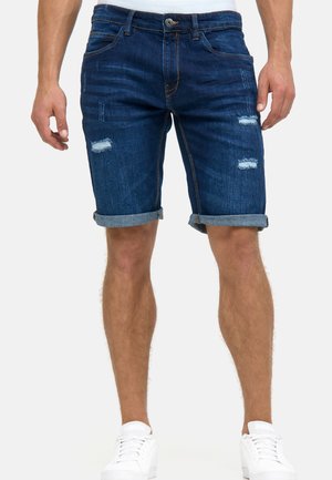Mann trägt dunkelblaue, hochgekrempelte Jeansshorts mit leichten Gebrauchsspuren an den Oberschenkeln und weiße Sneakers vor einfarbigem Hintergrund.