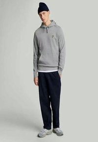 Hanorac gri cu glugă, decorat cu un mic logo galben, asortat cu pantaloni de trening negri și pantofi sport gri deschis. Purtați o căciulă groasă de tricotati navy.