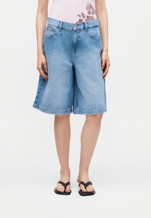 Žena u širokim svijetloplavim traper bermudama i crnim japankama stoji s rukama pokraj tijela.