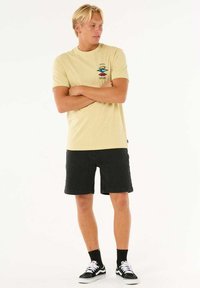 Lichtgele katoenen T-shirt met grafisch ontwerp, gecombineerd met zwarte shorts. Accessoires zijn zwarte sokken en zwarte sneakers.