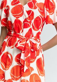 Robe rouge et blanche avec de grands motifs circulaires rouges abstraits, manches courtes, décolleté en V et une ceinture nouée à la taille pour sculpter.