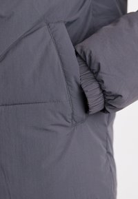 Doudoune grise en tissu doux et texturé, avec des poignets élastiques et une poche latérale ornée de détails de couture diagonale.