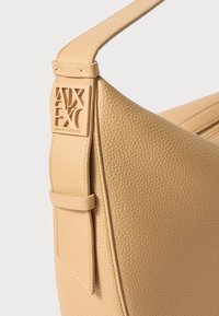 Borsa a tracolla in pelle beige con texture, dotata di una tracolla larga, con un'etichetta logo prominente e design minimalista. Dettagli cuciti sottili arricchiscono la forma.