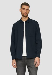 Camicia blu navy con bottoni, maniche lunghe, colletto, taschino frontale e una maglietta bianca sotto, abbinata a jeans grigio scuro.