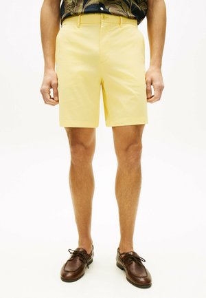 Mann trägt hellgelbe maßgeschneiderte Shorts, dunkelbraune Lederschuhe und ein gemustertes dunkles Hemd, steht vor weißem Hintergrund.