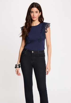 Femme aux longs cheveux foncés portant un haut bleu marine sans manches avec des détails en dentelle et un jean noir taille haute, accessoirisée de bracelets argentés.