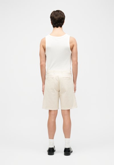 Άνδρας που στέκεται με την πλάτη στραμμένη, φοράει λευκό αμάνικο πουκάμισο, μπεζ σορτς, λευκές κάλτσες και μαύρα παπούτσια σε απλό φόντο.