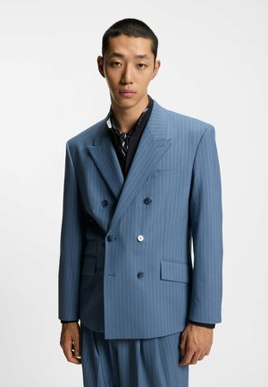 Abito doppiopetto azzurro chiaro con righe verticali sottili. Presenta rever a incavo, due tasche frontali e grandi bottoni. Tessuto strutturato.