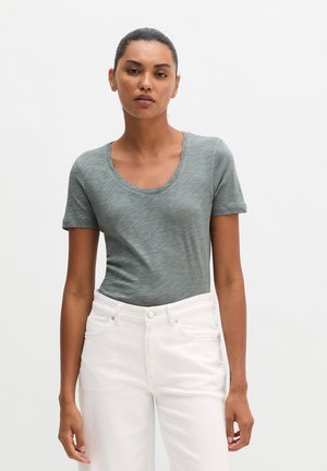Femme aux cheveux attachés portant un t-shirt gris à manches courtes et col rond ainsi qu’un jean blanc taille haute, debout devant un fond uni.