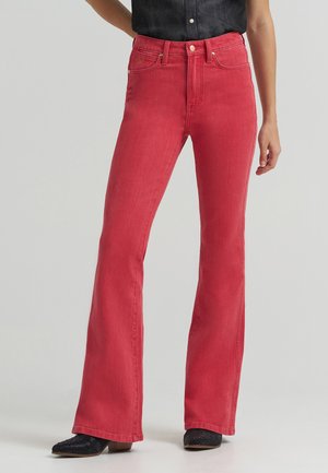 Frau trägt hoch taillierte, rote Jeans mit weitem Bein, kombiniert mit schwarzen Schuhen und einem dunklen ärmellosen Oberteil vor einfarbigem Hintergrund.