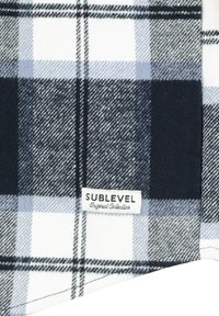 Kariertes Gewebe in Dunkelblau, Hellblau und Weiß. Weiche Textur mit einem Etikett mit der Aufschrift "SUBLEVEL Original Collection."