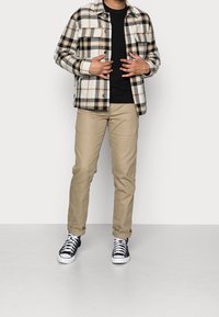 Chemise en flanelle à carreaux crème, noir et or, associée à un t-shirt noir et un chino beige. Des baskets noires et blanches complètent la tenue.