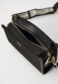 Borsa rettangolare nera con un motivo testurizzato, zip e hardware dorati. Include una tracolla in tessuto con dettagli del logo. Aperta per rivelare l'interno.