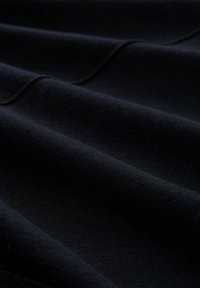 Close-up van donkere stof met zachte plooien en zichtbare stiksels langs de vouwen, die textuur en diepte tonen.