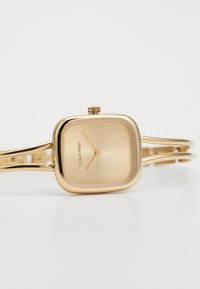 Gold square watch with a smooth dial, slim two-tone bracelet, minimal hour markers, and a textured crown. Engraved brand name visible.