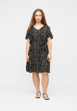 Kobieta stoi w czarnej sukience do kolan z białymi abstrakcyjnymi liniami, z krótkimi rękawami i wiązanym w talii paskiem, nosi czarne sandały.