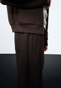 Bruin sweatshirt met ribbelmanchetten, gedragen over een grijze top. Donkerbruine broek met twee opgestikte zakken en scherpe plooien, gemaakt van zachte stof.